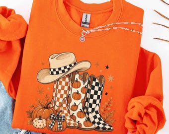 May include: A bright orange crewneck sweatshirt featuring a graphic print of a beige cowboy hat and two cowboy boots with checkered patterns. The design includes several orange pumpkins, autumn leaves, and small star shapes surrounding the boots.