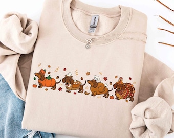 May include: A beige crewneck sweatshirt featuring a graphic print of four dachshunds dressed in autumn-themed costumes, including a pumpkin, a pie, a chef's hat, and a turkey, surrounded by small falling leaves and acorns.