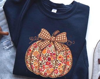 May include: A navy blue crewneck sweatshirt featuring a central graphic of a pumpkin. The pumpkin is filled with a vibrant floral pattern in shades of orange, red, and yellow, and is topped with a brown and white gingham patterned bow.