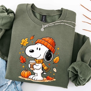May include: An olive green crewneck sweatshirt featuring a graphic of a white cartoon dog wearing an orange knit beanie and a matching striped scarf. The dog holds a coffee cup next to an orange pumpkin, surrounded by falling autumn leaves.