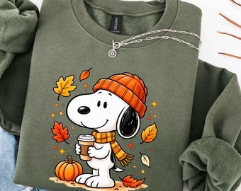 May include: An olive green crewneck sweatshirt featuring a graphic of a white cartoon dog wearing an orange knit beanie and a matching striped scarf. The dog holds a coffee cup next to an orange pumpkin, surrounded by falling autumn leaves.