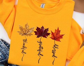 May include: A bright yellow crewneck sweatshirt featuring a graphic of three maple leaves in gold, deep red, and orange. Each leaf stem transitions into cursive text reading Thankful, Grateful, and Blessed in black ink.