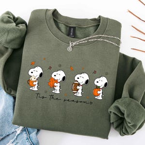 May include: An olive green crewneck sweatshirt featuring a graphic of Snoopy in four poses with autumn items including a mug, a leaf, a football, and a pumpkin. The text "Tis the season" is printed below the characters in a black script font.
