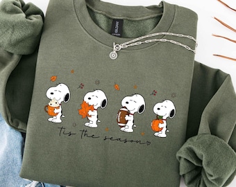 May include: An olive green crewneck sweatshirt featuring a graphic of Snoopy in four poses with autumn items including a mug, a leaf, a football, and a pumpkin. The text "Tis the season" is printed below the characters in a black script font.