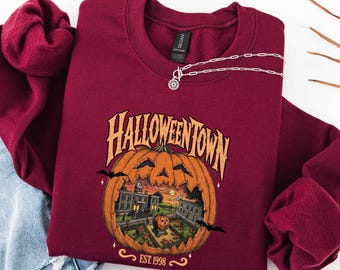 May include: A maroon crewneck sweatshirt featuring a graphic of a large carved pumpkin with a town scene inside. The text reads HALLOWEENTOWN and EST. 1998. A silver chain necklace with a circular pendant rests on the neckline of the garment.