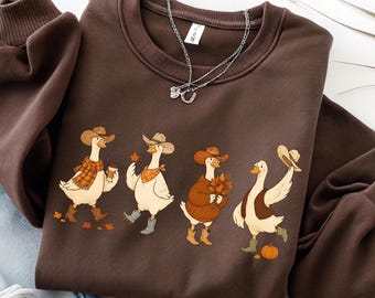 May include: A dark brown crewneck sweatshirt featuring a graphic print of four white geese wearing cowboy hats and boots. The geese are depicted with autumn-themed accessories including a plaid scarf, a coffee cup, fall leaves, and a pumpkin.
