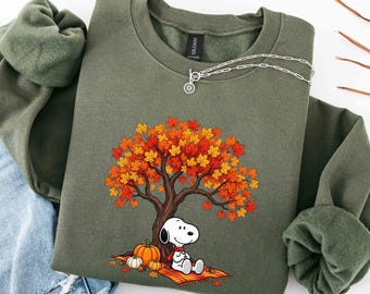 May include: An olive green crewneck sweatshirt featuring a graphic of Snoopy sitting under a tree with orange and yellow autumn leaves. The design includes a small orange pumpkin and a plaid blanket. A silver chain necklace rests on the collar.
