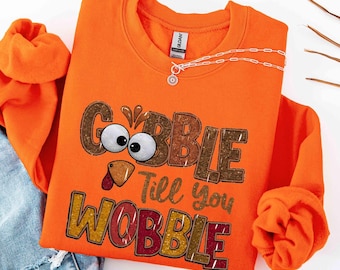 May include: An orange crewneck sweatshirt featuring a graphic of a turkey face with large white eyes and a brown beak. The text reads "GOBBLE Till you WOBBLE" in large, textured, multi-colored block letters. A silver chain necklace rests on the collar.