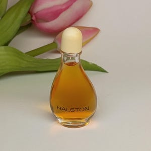 May include: A small, clear glass perfume bottle with a teardrop shape, filled with an amber-colored liquid. The bottle features a rounded, cream-colored cap and the word HALSTON printed in black lettering on the front of the glass surface.