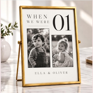 May include: A gold-toned metal picture frame containing a white print with the text "WHEN WE WERE 01" and "ELLA & OLIVER". The print features two black and white photographs of children, one showing a child in overalls and one showing a child in a dress.