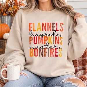 May include: A beige crewneck sweatshirt featuring the text FLANNELS, hayrides, PUMPKINS, sweaters, and BONFIRES printed in a distressed, multi-colored font with shades of red, orange, and yellow, set against a neutral fabric background.