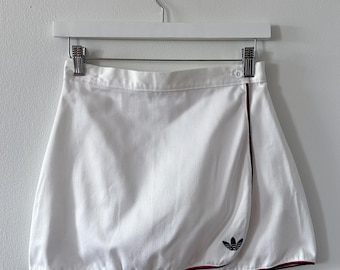 May include: A white wrap-style skirt featuring a high waistband with a single button closure. The hem and the overlapping front edge are accented with thin red and navy blue piping. A small navy blue trefoil logo is embroidered on the front.