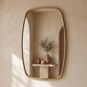 Op de afbeelding: Een wandspiegel met een afgerond rechthoekig frame in een metallic gouden afwerking. Het reflecterende oppervlak toont een stenen voetstuk met drie terracotta-kleurige keramische vazen en een groene tak in een neutrale kamer.