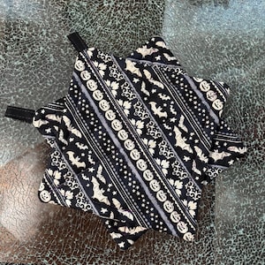 May include: Two square quilted pot holders featuring a black fabric base with a repeating pattern of white pumpkins, bats, stars, and ornate scrollwork. Each pot holder includes a black fabric hanging loop attached to one corner for storage.
