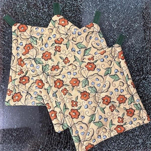 May include: Three square quilted pot holders featuring a cream background with a repeating pattern of orange flowers, green leaves, and small blue accents. Each pot holder includes a dark green fabric loop attached to one corner for hanging.