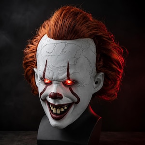 Puede incluir: Una máscara de payaso con rostro blanco de textura agrietada, nariz roja y marcas rojas que se extienden desde los ojos hasta la boca. La máscara tiene cabello rojo brillante, ojos rojos brillantes y una amplia sonrisa con dientes amarillentos.