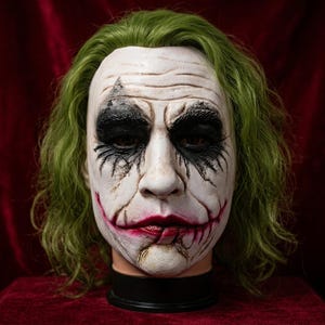 Puede incluir: Una máscara de cabeza completa con rostro blanco, maquillaje negro intenso en los ojos, una sonrisa roja pintada con cicatrices que se extienden desde las comisuras de la boca y cabello verde desordenado hasta los hombros sobre un soporte.