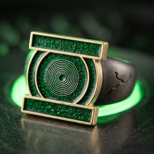 May include: A ring featuring a prominent rectangular and circular emblem with a textured green inlay and gold-toned metal borders. The band is dark gray with a matte finish and a subtle, etched line detail on the side of the metal shank.