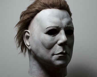 May include: A full-head mask featuring a pale, white, human-like face with deep-set black eye sockets, a neutral expression, and textured brown hair attached to the top and sides. The mask has defined facial features including a nose and lips.