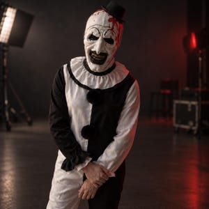 Könnte beinhalten: Ein Clownskostüm bestehend aus einem schwarz-weiß farblich abgesetzten Langarm-Oberteil mit Rüschenkragen und schwarzen Bommeln. Die Maske ist weiß mit schwarzen Augen- und Mundpartien, roten Blutspritzern und einem kleinen schwarzen Zylinder.
