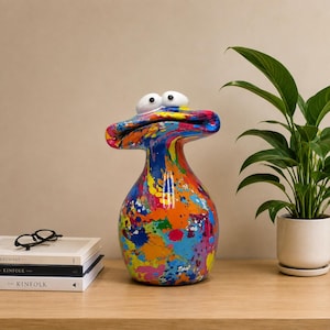 Peut inclure: Une sculpture décorative en céramique en forme de créature à large bouche avec deux grands yeux blancs. La surface présente un motif éclaboussé de peinture multicolore vif dans des tons de bleu, orange, jaune, rose et vert avec une finition brillante.