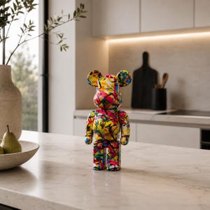 Peut inclure: Une figurine en forme d'ours arborant un motif graffiti multicolore vif avec des éclaboussures de jaune, rose, bleu et noir. La figurine possède des membres articulés, des oreilles arrondies et une finition lisse et brillante, posée debout.