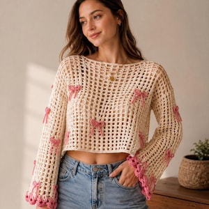 May include: A cream-colored, long-sleeved, open-knit crochet sweater featuring a wide boat neckline and a cropped hem. The sweater is decorated with small, pink, three-dimensional crochet bows scattered across the front and sleeves, with pink scalloped trim at the cuffs.