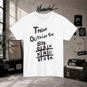 Può includere: Una t-shirt bianca a maniche corte con il testo nero "THINK OUTSIDE THE BOX" sopra una griglia di tris disegnata a mano. La griglia contiene segni neri a forma di X e O, con una linea rossa verticale tracciata attraverso la colonna centrale.