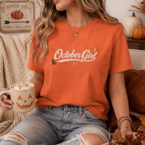 May include: An orange short-sleeve t-shirt featuring the white text October Girl, a small black cat graphic, a crescent moon, and a pumpkin icon. The shirt has a crew neckline and is worn with light blue denim jeans that have distressed knees.