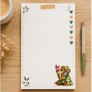May include: A rectangular notepad with a white grid pattern, featuring a dashed border, green leaf illustrations in the corners, and a column of alternating green and tan hearts. A small illustration of a bear and a dragon eating ice cream is included.