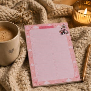 Puede incluir: Un bloc de notas rectangular de color rosa con una doble columna de casillas de verificación y líneas para escribir. El borde está decorado con nubes blancas, corazones pequeños, estrellas e ilustraciones de algodón de azúcar. El texto EINKAUFSLISTE aparece arriba.