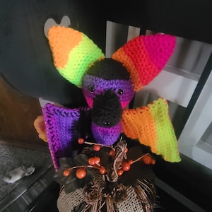 May include: A crocheted rainbow bat with black eyes and a purple and black body. The bat is sitting on a brown and burlap pumpkin with orange berries.