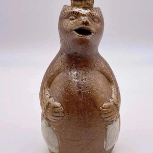 May include: A brown ceramic bottle shaped like a standing bear with a textured surface, a wide open mouth, and two light-colored pockets on the front. The top of the bear's head features a small cork stopper inserted into the opening of the vessel.