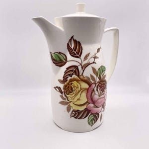 May include: A cream-colored ceramic coffee pot featuring a crackle glaze finish and a floral design. The illustration displays large yellow and pink roses surrounded by brown and green leaves. It includes a matching lid, a handle, and a spout.