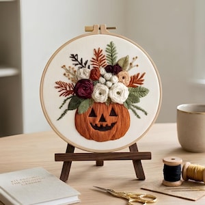 May include: A circular wooden embroidery hoop containing a fabric panel with a hand-stitched design of a jack-o'-lantern pumpkin overflowing with a bouquet of white, burgundy, and orange flowers and green foliage, displayed on a small wooden easel.