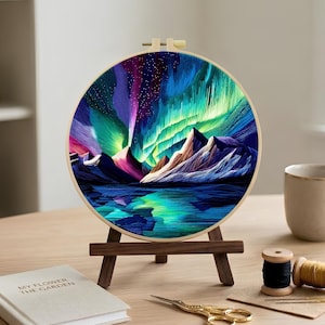 May include: A circular embroidery hoop featuring a vibrant needlework depiction of the aurora borealis above jagged mountain peaks and a reflective body of water, displayed on a small wooden easel stand with a light wood frame and tension screw.