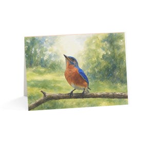 May include: A folded greeting card featuring a watercolor painting of a bluebird with a bright blue head and wings and a rusty orange chest. The bird is perched on a thin brown branch against a soft, blurred green forest background.