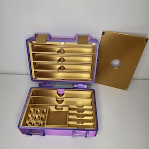 Peut inclure: Une mallette de rangement rectangulaire avec un extérieur violet métallique et un intérieur doré. La mallette est ouverte, révélant plusieurs compartiments, dont des fentes horizontales, une section carrée et de petites alvéoles circulaires. Un couvercle doré séparé est posé à droite.