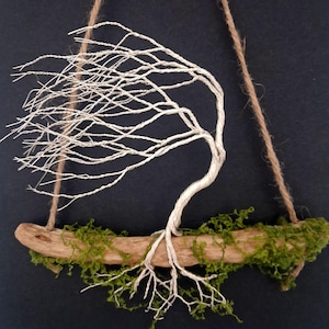 May include: A wall hanging featuring a silver wire tree sculpture attached to a piece of driftwood covered in green moss. The tree branches are shaped as if blowing in the wind, and the entire piece is suspended by a brown jute twine cord.
