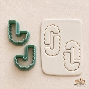 May include: Two sage green plastic cookie cutters shaped like the letter J with a scalloped edge, alongside a rectangular piece of white rolled dough featuring two identical J-shaped impressions. The text THE CROWN COLLECTIVE appears at the bottom.