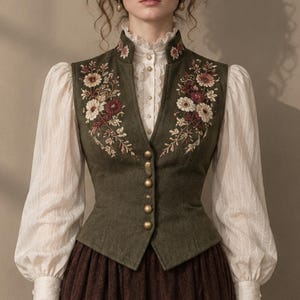 Può includere: Un gilet verde oliva con ricami floreali nei toni del rosso, bianco e oro, caratterizzato da un colletto alto e bottoni color ottone. È indossato sopra una camicetta color crema a maniche lunghe con colletto e polsini arricciati e una gonna marrone.