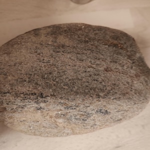 Può includere: Una pietra di fiume liscia di forma ovale con una consistenza maculata caratterizzata da una miscela di granelli minerali grigi, marroni e neri. La superficie mostra variazioni naturali di colore e grana, creando un aspetto punteggiato su tutta la roccia.