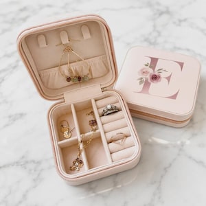 Puede incluir: Un joyero cuadrado de color rosa claro con cierre de cremallera. La tapa presenta una letra E grande con un diseño floral. El interior está forrado con tela beige e incluye compartimentos para anillos, pendientes y collares con joyas de oro.