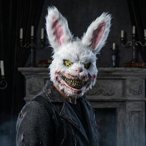 Può includere: Una maschera da coniglio in pelliccia sintetica bianca con lunghe orecchie dai dettagli interni rosa, occhi gialli con pupille nere a forma di croce e un ampio sorriso minaccioso che mostra denti giallastri e macchie rosso sangue.