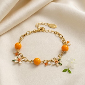 Puede incluir: Una pulsera de cadena en tono dorado con dos cuentas redondas de color naranja y dos dijes de flores de esmalte blanco con hojas verdes. La pulsera incluye pequeñas cuentas de acento naranjas y blancas, un cierre de langosta y una etiqueta ovalada.