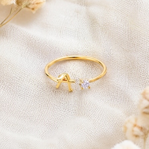 Puede incluir: Un anillo abierto de tono dorado que presenta una letra A mayúscula en un extremo y una pequeña piedra preciosa redonda y transparente en el otro. La banda es fina y circular, diseñada con un espacio ajustable entre la letra y la piedra.