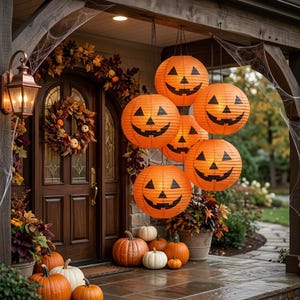 Op de afbeelding: Een verzameling oranje papieren lantaarns in de vorm van pompoenen met zwarte gezichten die aan een veranda hangen. De veranda is versierd met een herfstkrans, herfstbladeren en diverse pompoenen die op de stenen vloer zijn geplaatst.