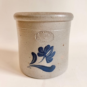 May include: A cylindrical gray stoneware crock with a speckled texture and a thick rim. It features a blue painted floral design and an oval stamp that reads Rockdale Union Stoneware 1924 Always Handmade.