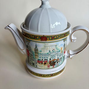 Puede incluir: Una tetera de cerámica blanca con tapa abovedada y borde dorado en el asa y el pico. El cuerpo presenta una ilustración colorida de monumentos de Londres, incluyendo el Big Ben y un carruaje real, con un borde decorativo en verde y oro.