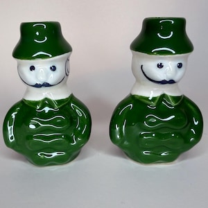 May include: A pair of ceramic salt and pepper shakers shaped like figures wearing green hats and green jackets with white collars. The figures have white faces, dark blue painted eyes, and dark blue mustaches. The glossy finish reflects light.